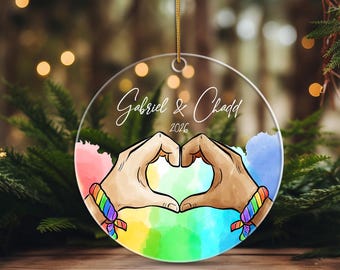 Può includere: Un ornamento rotondo in acrilico trasparente con un'illustrazione stampata di due mani che formano un cuore. I polsi sono decorati con braccialetti color arcobaleno. Il testo Gabriel & Chadd e l'anno 2026 sono stampati in corsivo bianco.