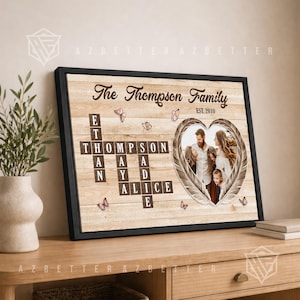 May include: A framed wall art print featuring a light wood grain background with the text The Thompson Family EST. 2010. It includes a crossword-style layout of names Ethan, Thompson, May, Sadie, and Alice, plus a heart-shaped photo frame with butterflies.