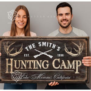 May include: A rustic wooden sign with the text "THE SMITH'S HUNTING CAMP" featuring crossed rifles, antlers, and the year 1990. The sign also includes the location "Lake Morena, California".