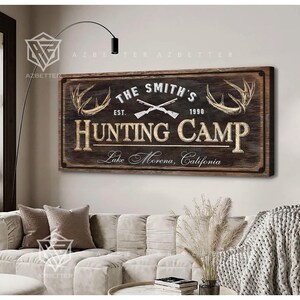 May include: A rustic wooden sign that reads "The Smith's Hunting Camp" with crossed rifles, antlers, and the text "Lake Morena, California." The sign is brown with cream-colored lettering and is mounted on a wall above a sofa.