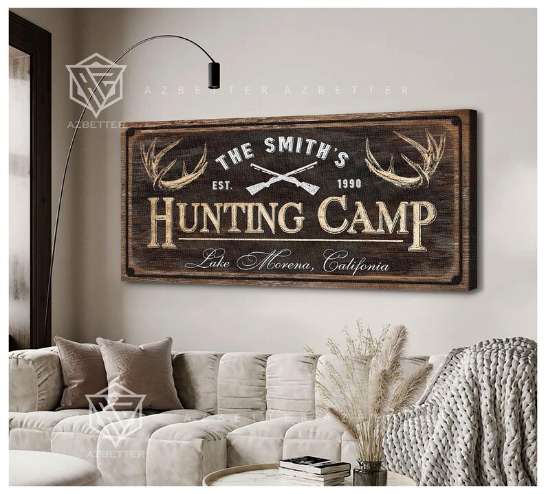 May include: A rustic wooden sign that reads "The Smith's Hunting Camp" with crossed rifles, antlers, and the text "Lake Morena, California." The sign is brown with cream-colored lettering and is mounted on a wall above a sofa.