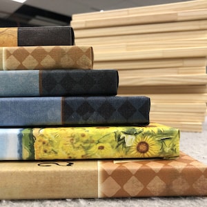 May include: A stack of five canvas prints with various designs. The top print features a yellow sunflower design, the second print has a blue and white sky design, the third print has a blue and black design, the fourth print has a brown and white diamond pattern, and the bottom print has a yellow and brown design.