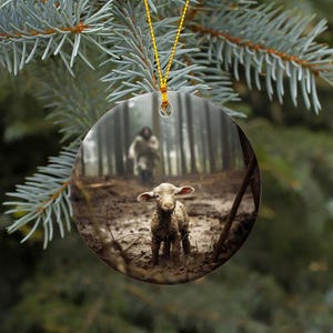 Pu&ograve; includere: Un ornamento natalizio rotondo con un agnello bianco in piedi in una radura boscosa fangosa. Una figura sullo sfondo cammina verso l'agnello.