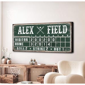 May include: A green and white baseball scoreboard with the text "ALEX FIELD". The scoreboard displays the scores for visitor and home teams, with the text "BALL 2", "STRIKE 2", and "OUT 1".