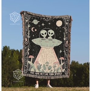 May include: Black woven throw blanket featuring a UFO with two aliens, the names "Henry" and "Olivia", and the text "YOU ARE OUT OF THIS WORLD". The blanket has a fringe border and a space-themed design with mushrooms and planets.