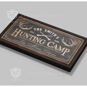 May include: A rectangular wooden sign with a dark brown frame. The sign reads "THE SMITH'S HUNTING CAMP" with crossed rifles and antlers. The text "Lake Morena, California" is also included. The sign has a rustic, vintage aesthetic.