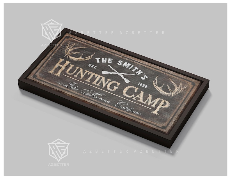 May include: A rectangular wooden sign with a dark brown frame. The sign reads "THE SMITH'S HUNTING CAMP" with crossed rifles and antlers. The text "Lake Morena, California" is also included. The sign has a rustic, vintage aesthetic.