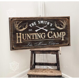 May include: A rectangular wooden sign with the text "THE SMITH'S HUNTING CAMP" and "Lake Morena, California." The sign features crossed rifles, antlers, and the year "1990." The sign is brown with white and gold lettering.
