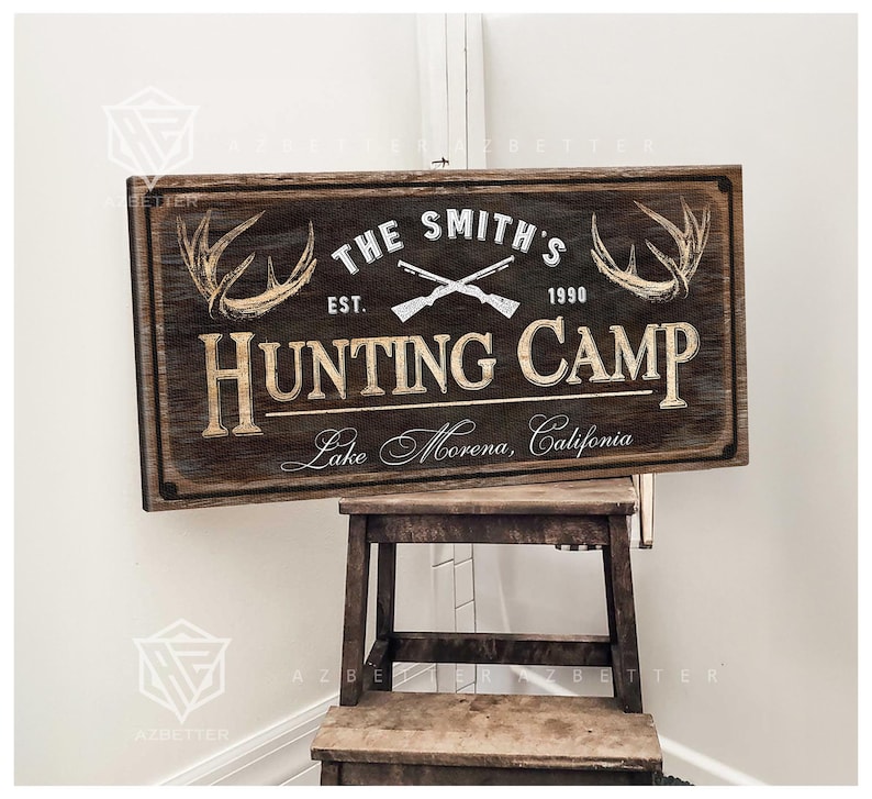 May include: A rectangular wooden sign with the text "THE SMITH'S HUNTING CAMP" and "Lake Morena, California." The sign features crossed rifles, antlers, and the year "1990." The sign is brown with white and gold lettering.