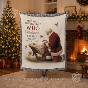 May include: A woven throw blanket featuring a cream background with a fringed edge. The design depicts Santa Claus kneeling beside a manger with a baby, a small lamb, and birds. The text reads: MAY WE NEVER FORGET WHO CHRISTMAS IS REALLY ABOUT.