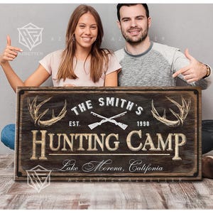 May include: A rustic wooden sign with the text "THE SMITH'S HUNTING CAMP" featuring crossed rifles, antlers, and the establishment year 1990. The sign also includes the location "Lake Morena, California".
