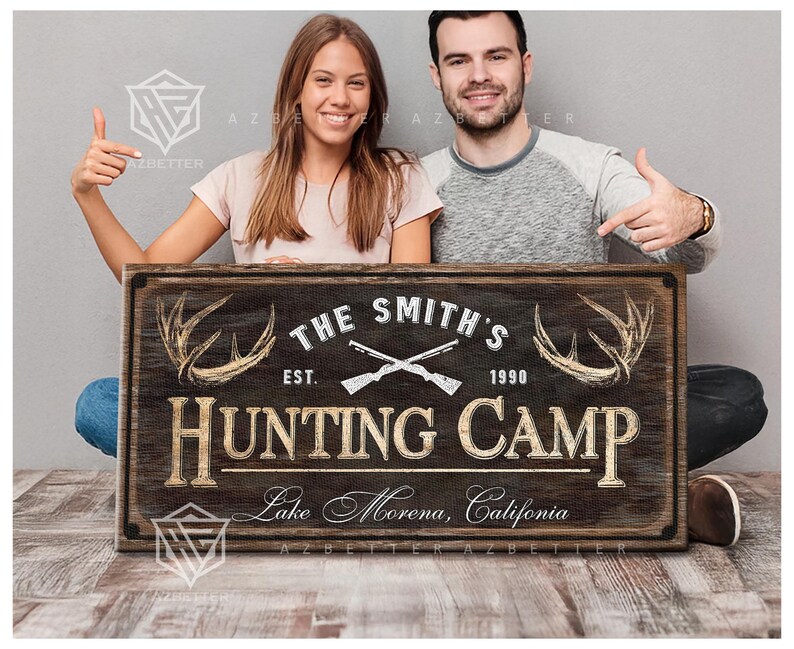 May include: A rustic wooden sign with the text "THE SMITH'S HUNTING CAMP" featuring crossed rifles, antlers, and the establishment year 1990. The sign also includes the location "Lake Morena, California".