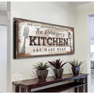 Könnte beinhalten: Ein rechteckiges Holzschild mit dem Text "The Robinson's KITCHEN ARE MADE HERE" und dem Jahr 2024. Das Schild hat ein rustikales Design mit einer Gabel- und Löffelillustration. Das Schild hängt über einer dunkelbraunen Holzkonsolentisch mit drei Topfpflanzen.