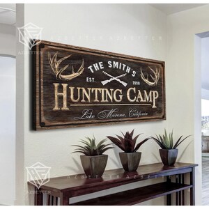 May include: A rustic wooden sign with the text "THE SMITH'S HUNTING CAMP" and the year "1990". The sign features crossed rifles and deer antlers. Below the sign are three potted plants on a dark wooden table.