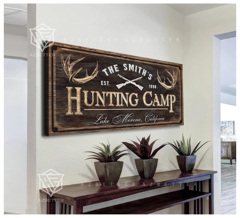May include: A rustic wooden sign with the text "THE SMITH'S HUNTING CAMP" and the year "1990". The sign features crossed rifles and deer antlers. Below the sign are three potted plants on a dark wooden table.