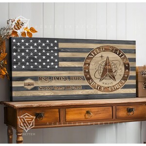 May include: Wooden American flag with the United States Space Force emblem. The flag design includes the text "MSgt. JAMES L. HAYES" and "Service Years: 1995-2020". The flag is displayed on a wooden table.