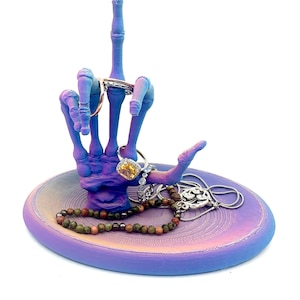 May include: A purple and blue iridescent skeleton hand ring holder with a matching dish. The hand is giving the middle finger and has several rings on it. The dish has a variety of jewelry on it, including a necklace, bracelets, and rings.