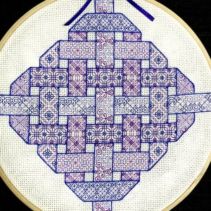 Puede incluir: Un aro de bordado circular con fondo blanco y un diseño geométrico azul y morado. El diseño es un patrón repetitivo de cuadrados y rectángulos, creando un efecto tejido.