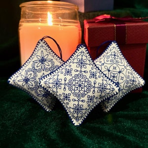 May include: Three white and blue embroidered diamond-shaped ornaments with intricate snowflake designs. A lit candle in a glass jar and a red gift box with a ribbon are in the background. The ornaments are hanging by blue loops.