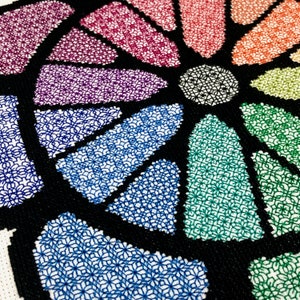 May include: A colourful cross-stitch design featuring a circular pattern of geometric shapes in shades of pink, red, orange, yellow, green, blue, purple, and black.