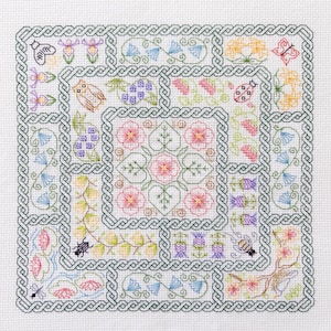 Op de afbeelding: Een vierkant kruissteekontwerp met een centraal bloemmotief omgeven door diverse geborduurde motieven. Het ontwerp bevat bloemen, insecten en een rand met een Keltisch knoopmotief. Het kleurenpalet omvat roze, groen, geel en blauw.