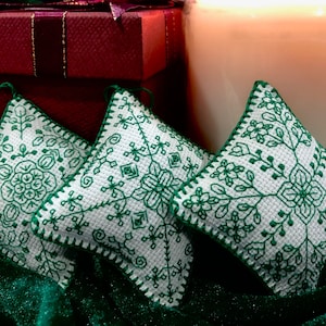 May include: Three white fabric ornaments with green embroidered designs. The ornaments are sitting on a green velvet surface.
