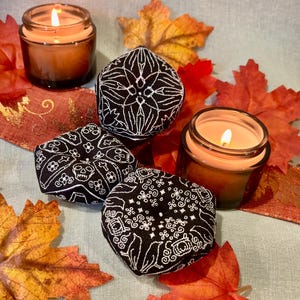 May include: Two lit candles in glass jars and three black fabric decorative objects with white embroidered designs. The objects have floral, geometric, and skull patterns. The scene is decorated with autumn leaves and a ribbon.