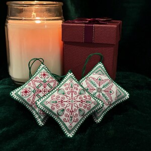 May include: Three white fabric ornaments with green and red embroidery designs. The ornaments are hanging from a green velvet surface. A lit candle and a red gift box are in the background.