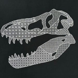 May include: White embroidered dinosaur skull on a black background. The skull is made up of a repeating geometric pattern.