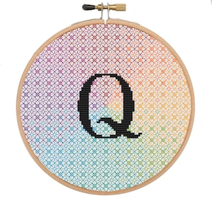 May include: A cross-stitch embroidery hoop featuring a gradient background of rainbow colors. The design includes a black letter "Q" in the center. The wooden hoop is secured with a brass screw.