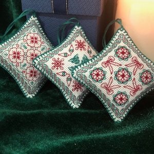 Könnte beinhalten: Drei weiße und grüne bestickte Kissenornamente mit roten Akzenten. Die Ornamente zeigen verschiedene festliche Designs, darunter Schneeflocken, Schleifen und Blumen.