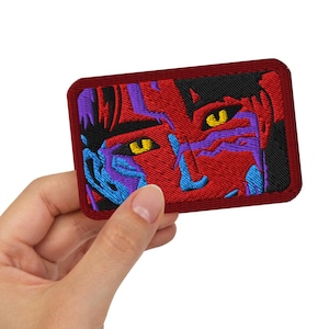 May include: Embroidered patch featuring a stylized face with yellow eyes, blue and purple accents, and a red background. The patch has a rectangular shape with rounded corners and a red border. The design is detailed and colorful.
