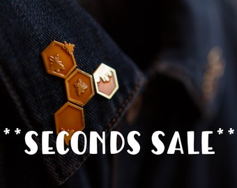 May include: Gold-colored bee-themed enamel pins in a honeycomb design, attached to a dark denim jacket. The pins feature bee and honey details. The text "SECONDS SALE" is displayed in white font.