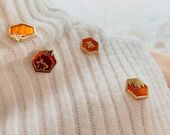 May include: Four gold-colored pins shaped like honeycombs with a bee design. The pins are on a white knitted sweater.