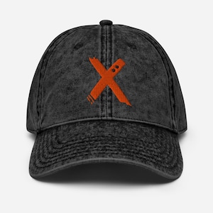 Può includere: Un cappellino da baseball nero in stile vintage con un design a "X" arancione ricamato. Il cappellino ha una visiera curva e una chiusura con cinturino in tessuto. La "X" ha un aspetto invecchiato, che aggiunge all'estetica vintage del cappellino.