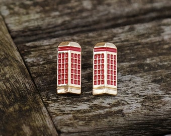 Puede incluir: Un par de pendientes dorados con forma de cabinas telefónicas rojas. Los pendientes tienen forma rectangular con cuerpo rojo, ribetes blancos y parte superior dorada. Se encuentran sobre un fondo de madera rústica.