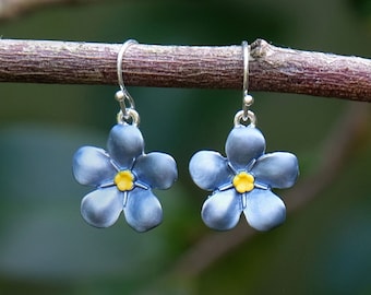 May include: A pair of silver-tone earrings featuring blue flower pendants with yellow centers. The earrings are hanging from a brown branch against a blurred green background. The flower design is detailed and delicate.