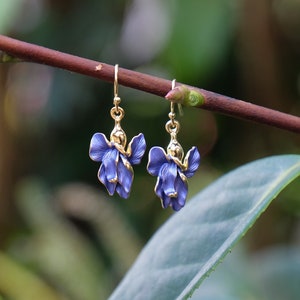 Puede incluir: Un par de pendientes dorados con un diseño de flor azul. Los pendientes cuelgan de una rama marrón con hojas verdes en el fondo. Los pendientes tienen una forma floral detallada y tridimensional.