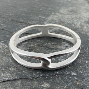 Puede incluir: Un brazalete de plata con un diseño simple y moderno. El brazalete está hecho de dos bandas que están entrelazadas y tienen un acabado liso y pulido.