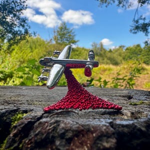 Op de afbeelding: Een glanzend zilveren vliegtuigmodel met vier motoren, gemonteerd op een levendige rode, getextureerde basis. Het model staat tegen een achtergrond van groen gebladerte en een blauwe lucht, wat een opvallend contrast creëert.