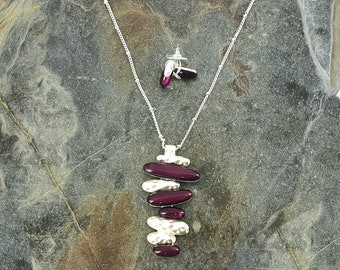 May include: A silver necklace with a pendant made of stacked, hammered silver and purple glass ovals. Matching earrings with purple glass ovals are also included.
