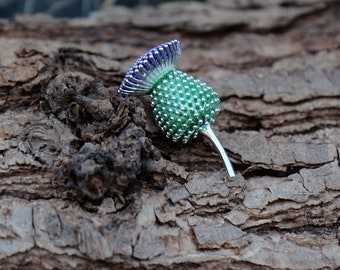 Puede incluir: Broche de cardo plateado con una cabeza verde texturizada y una parte superior morada y azul. El broche se encuentra sobre un fondo de madera natural, resaltando su intrincado diseño y detalle. Un accesorio decorativo.