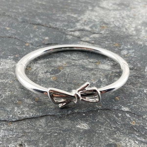 Puede incluir: Un brazalete de plata con un diseño de lazo. El brazalete está hecho de metal pulido y tiene un aspecto simple y elegante.