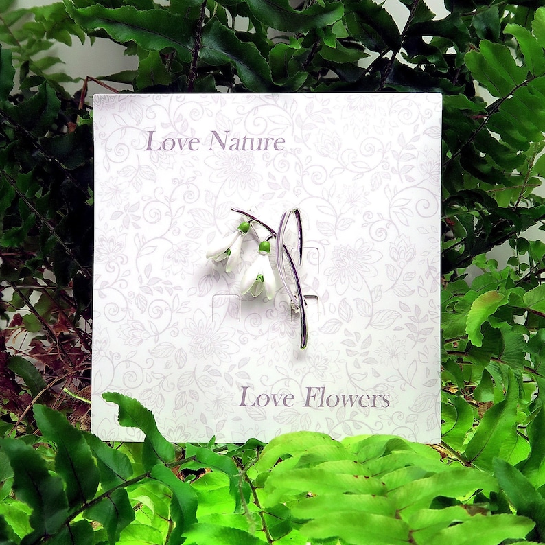 May include: A silver-toned brooch featuring a snowdrop flower design with white petals and green accents. The brooch is displayed on a white card with the text "Love Nature" and "Love Flowers" against a floral pattern, surrounded by green foliage.