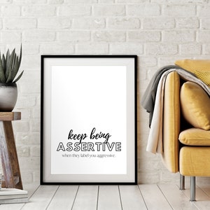 May include: A black framed print with the text "keep being assertive when they label you aggressive." The print is hanging on a white brick wall, with a yellow couch and plants in the foreground.
