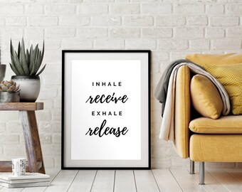 May include: A black framed print with a white background and the text "INHALE receive EXHALE release" in black cursive font. The print is hanging on a white brick wall above a yellow sofa.