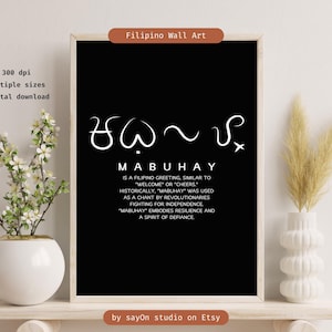 May include: A black framed print featuring the Filipino greeting "MABUHAY" in white script. The text explains the greeting's meaning and historical significance. The artwork is displayed on a shelf, accompanied by a white vase and floral arrangement.