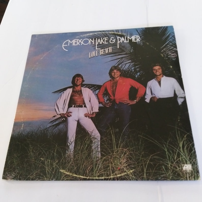 May include: A vintage vinyl record album cover featuring Emerson, Lake & Palmer. The cover art shows the band members standing in front of a tropical beach scene with palm trees and a sunset sky. The album title "Love Beach" is printed in white letters on a black background.