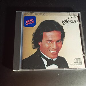 May include: A compact disc case featuring Julio Iglesias. The cover has a gold background with a portrait of Iglesias in a tuxedo. The text "Julio Iglesias" is displayed, along with a "Best Value" sticker.