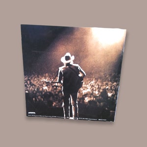 May include: A black and white photo of a country music singer performing on stage in front of a large crowd. The singer is wearing a cowboy hat and a long-sleeved shirt. The stage is lit with spotlights.